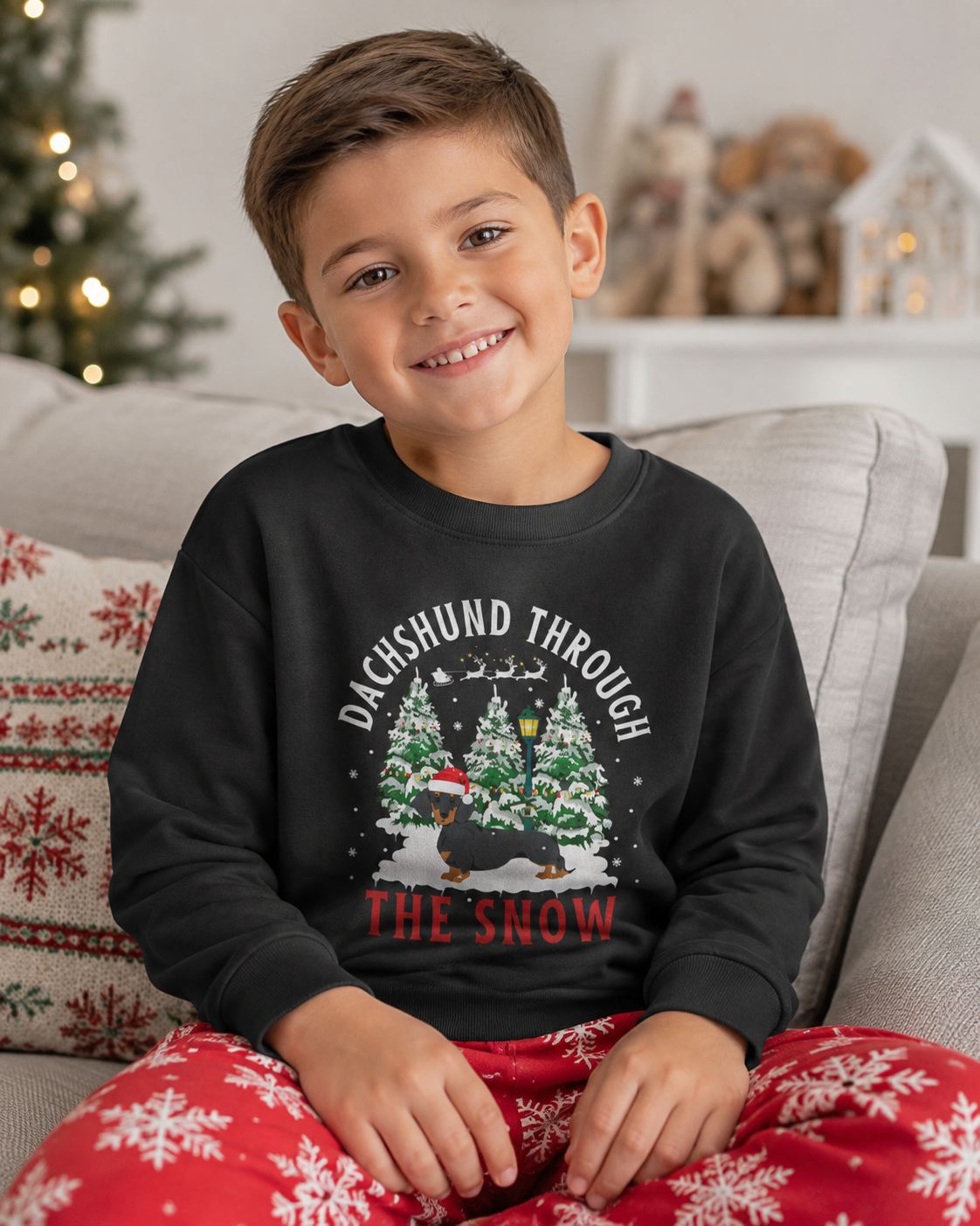 A young boy smiling while wearing a black crewneck sweatshirt with a 'Dachshund Through The Snow' Christmas graphic, paired with red holiday pajama pants with white snowflakes, sitting on a grey couch.