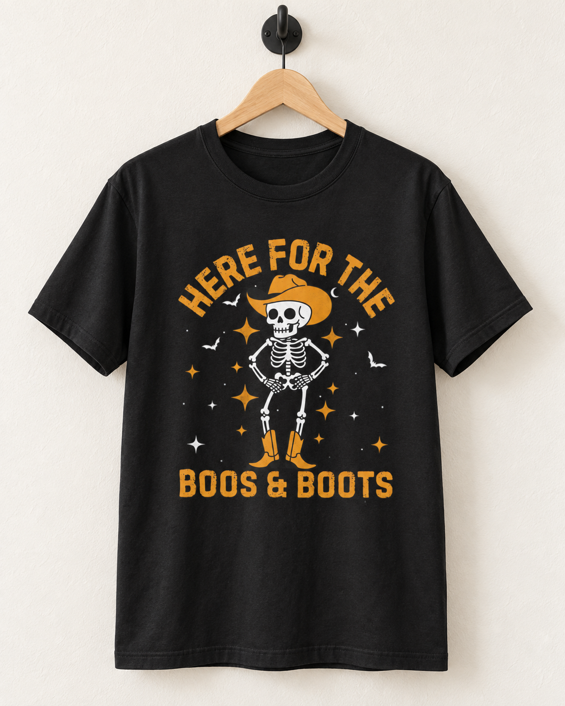 Black “Here for the Boos & Boots” cowboy skeleton T-shirt displayed on a wooden hanger and black wall hook.