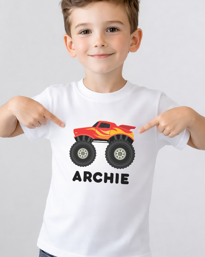 Young boy pointing at personalised white monster truck t-shirt featuring red flame truck graphic and name Archie in black text