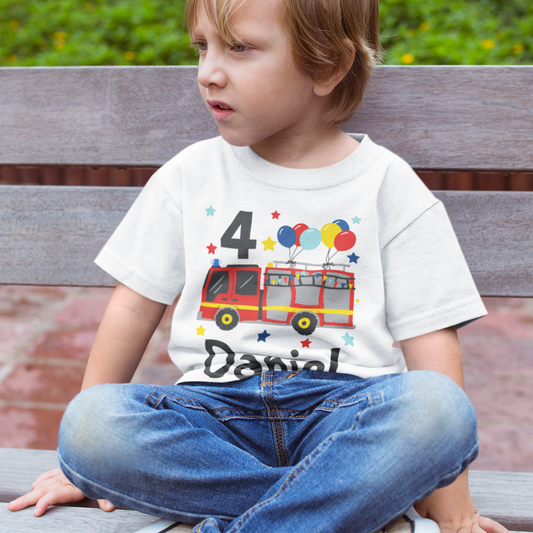 A little boy wearing a white t-shirt with a fire truck, bunting, balloons, stars design and the number 4, sitting on a wooden bench outdoors.