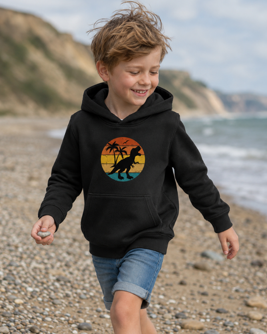 Boy walking on a windy beach wearing a black kids hoodie with a retro sunset T-rex dinosaur graphic, styled with denim shorts for a Jurassic Coast inspired look.