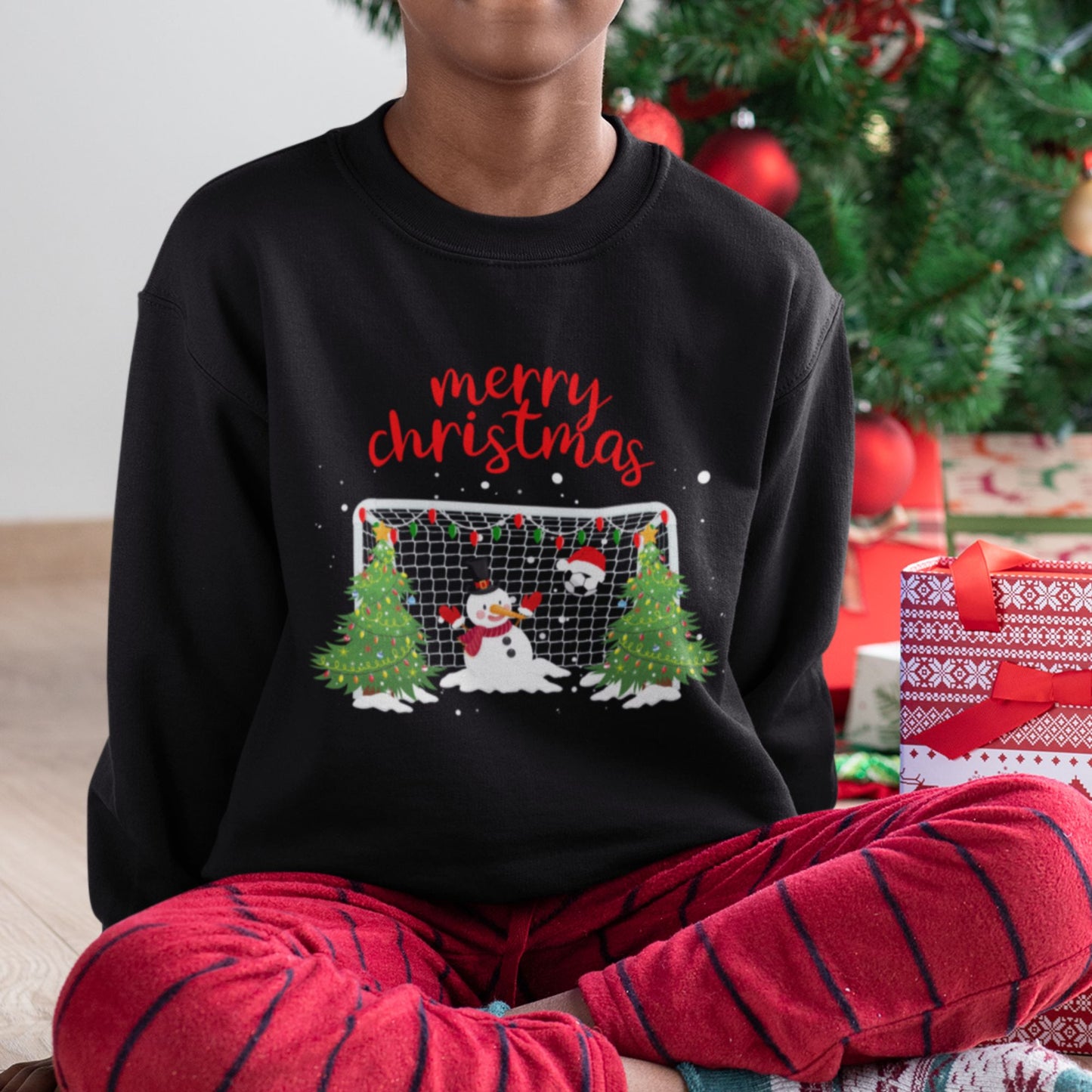 Boy wearing black Christmas sweatshirt with snowman goalkeeper and football goal design, festive Christmas trees, and “Merry Christmas” text.