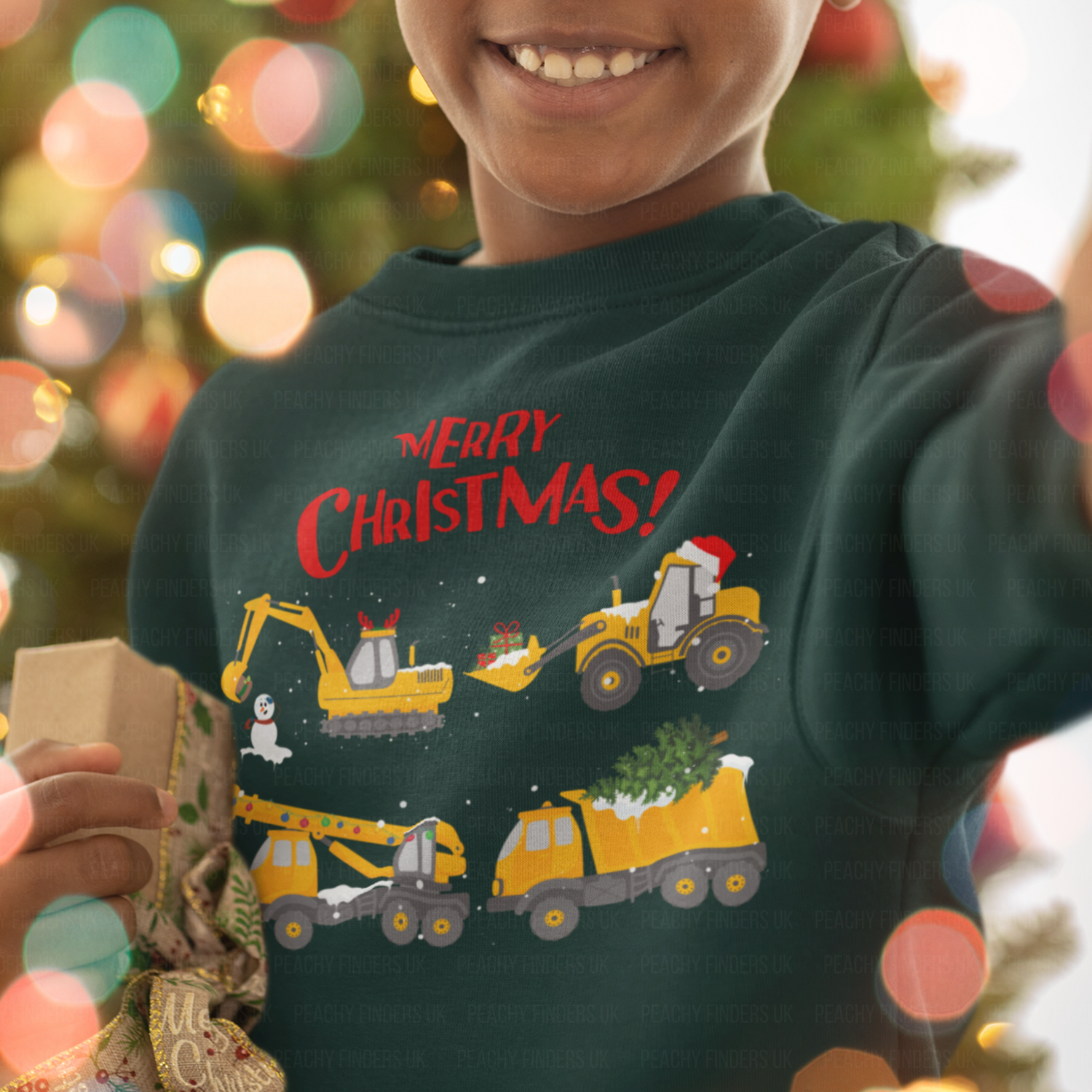 A young boy wearing a dark green Christmas sweatshirt featuring festive construction vehicles, including diggers and trucks carrying presents and a Christmas tree, with “Merry Christmas!” text on top.