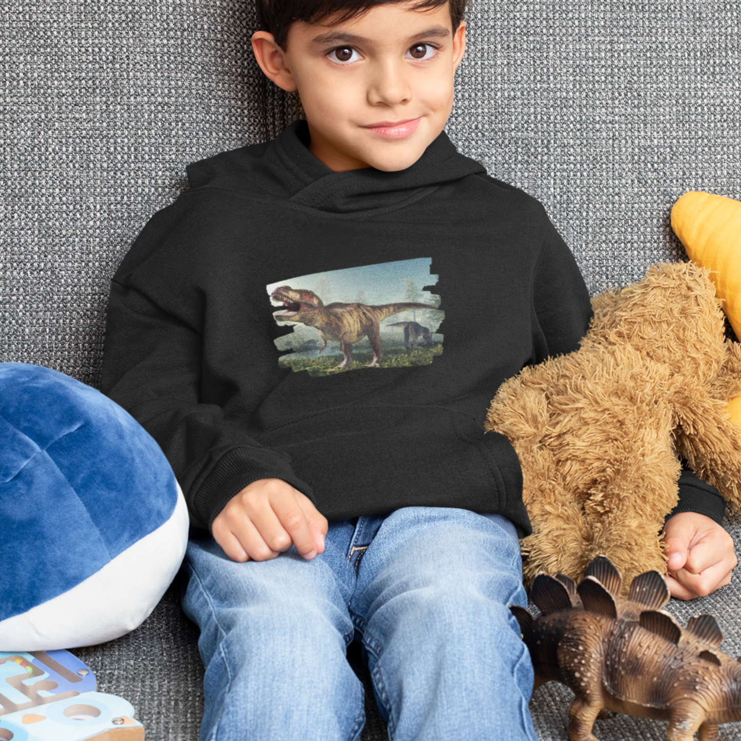 A boy wearing a black hoodie with a dinosaur graphic, sitting on a grey couch.