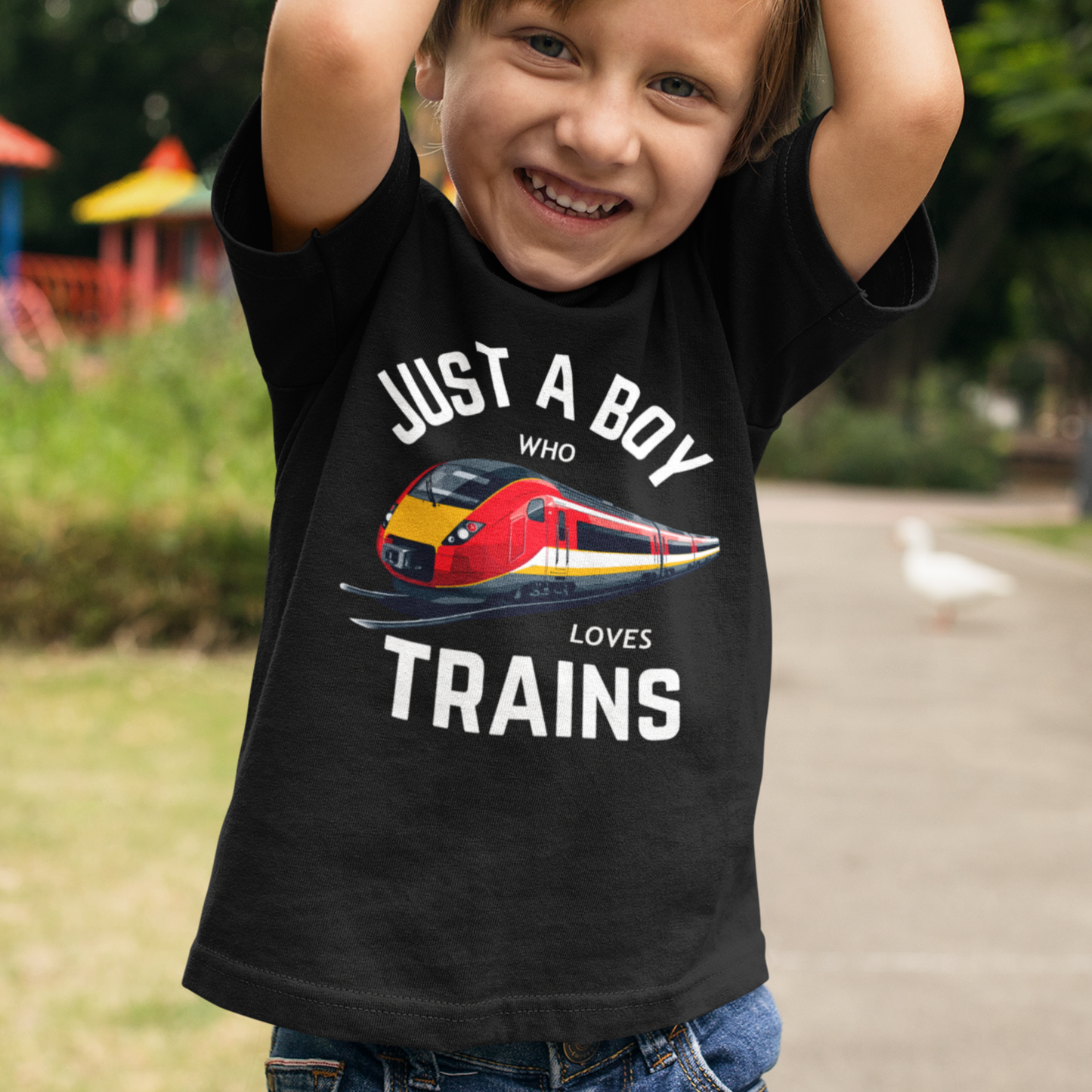 Smiling young boy wearing a black ‘Just a Boy Who Loves Trains’ T-shirt with a bright red and yellow train graphic, standing outdoors in a park.