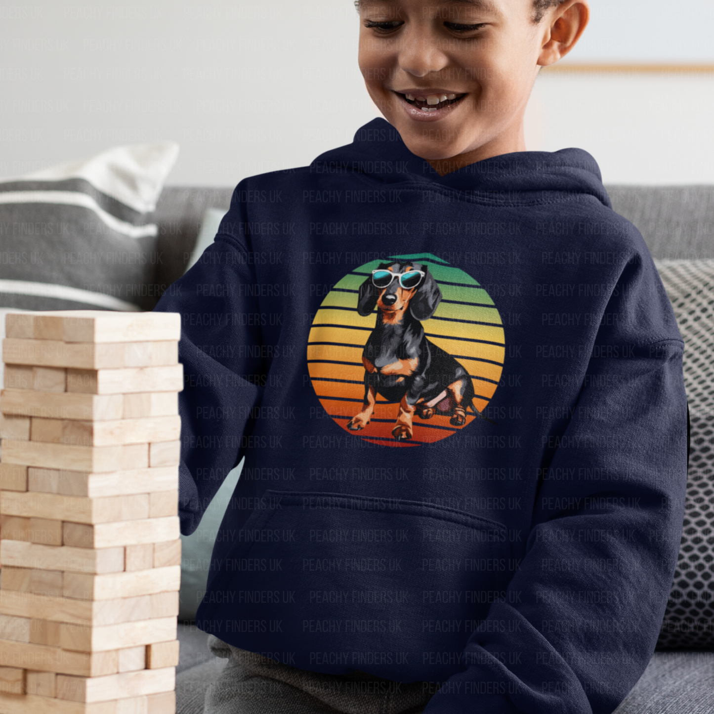 A smiling young boy with brown skin and short curly hair wears a navy blue hooded sweatshirt. The hoodie features a graphic of a black and tan Dachshund dog wearing sunglasses against a retro striped circle of orange, yellow, and green.