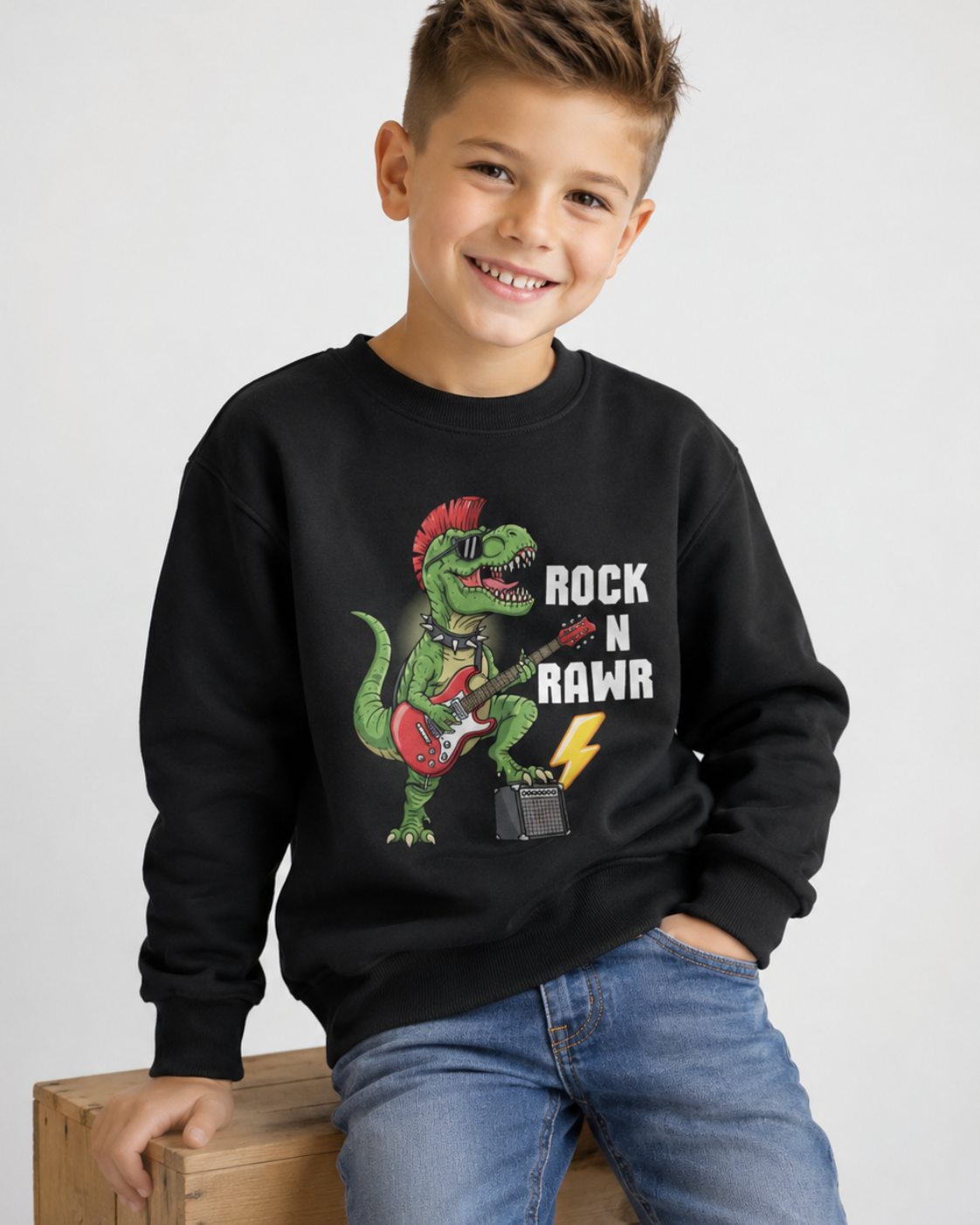A smiling young boy wearing a black crewneck sweatshirt with a colourful graphic of a green dinosaur playing a red guitar and the text 'ROCK N RAWR', paired with blue jeans, sitting on a wooden crate against a light grey background.