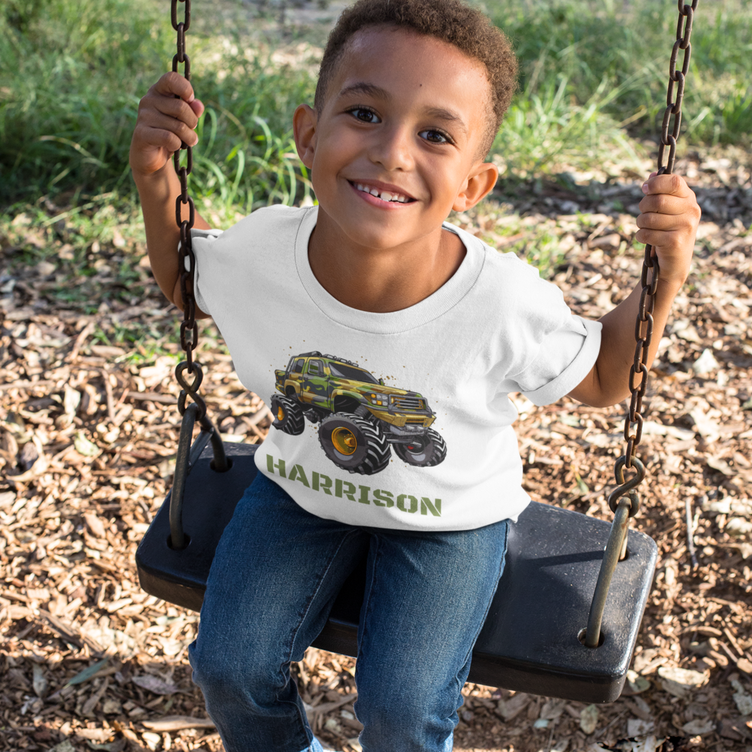 A child sitting on a swing wearing a white t-shirt with a camo monster truck design and the name 'Harrison' printed on the front.