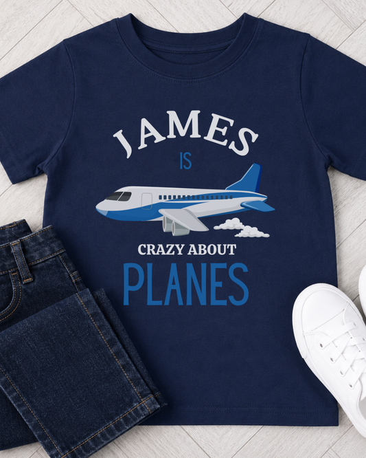 A navy blue kids' t-shirt with a custom graphic print reading 'JAMES IS CRAZY ABOUT PLANES' around an illustration of a blue and white airplane. The shirt is styled in a flat-lay with dark blue jeans, a white sneaker, and a light wood floor background.