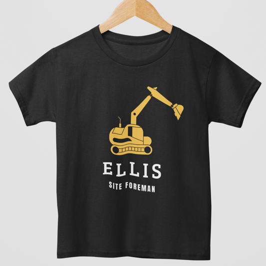 Kids black cotton t-shirt with a yellow excavator graphic and personalised name with “Site Foreman” text, styled on a wooden hanger.
