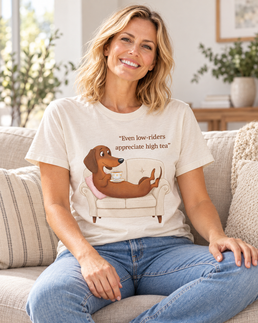 A blonde woman sitting cross-legged on a cream sofa wearing a natural-coloured short-sleeve t-shirt. The tee features an illustration of a brown dachshund lounging on an armchair holding a teacup, with the text "Even low-riders appreciate high tea" printed above.tea'.
