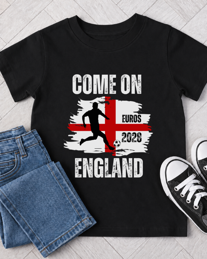 Kids Come On England Euros 2028 football T-shirt flat lay with St George’s flag design, footballer graphic, jeans and black trainers.