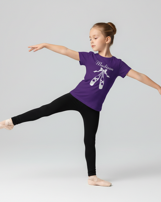 Young girl wearing a personalised purple ballet t-shirt with ballet shoes graphic and name print, paired with black leggings and ballet flats, smiling at the camera while posing in a ballet stance against a light studio background.