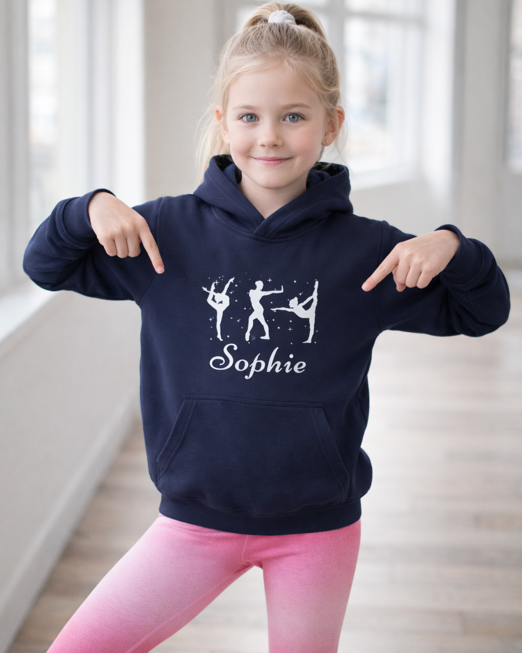 Girl pointing to the personalised name on a navy gymnastics hoodie featuring white gymnast silhouettes and Sophie text in a studio.