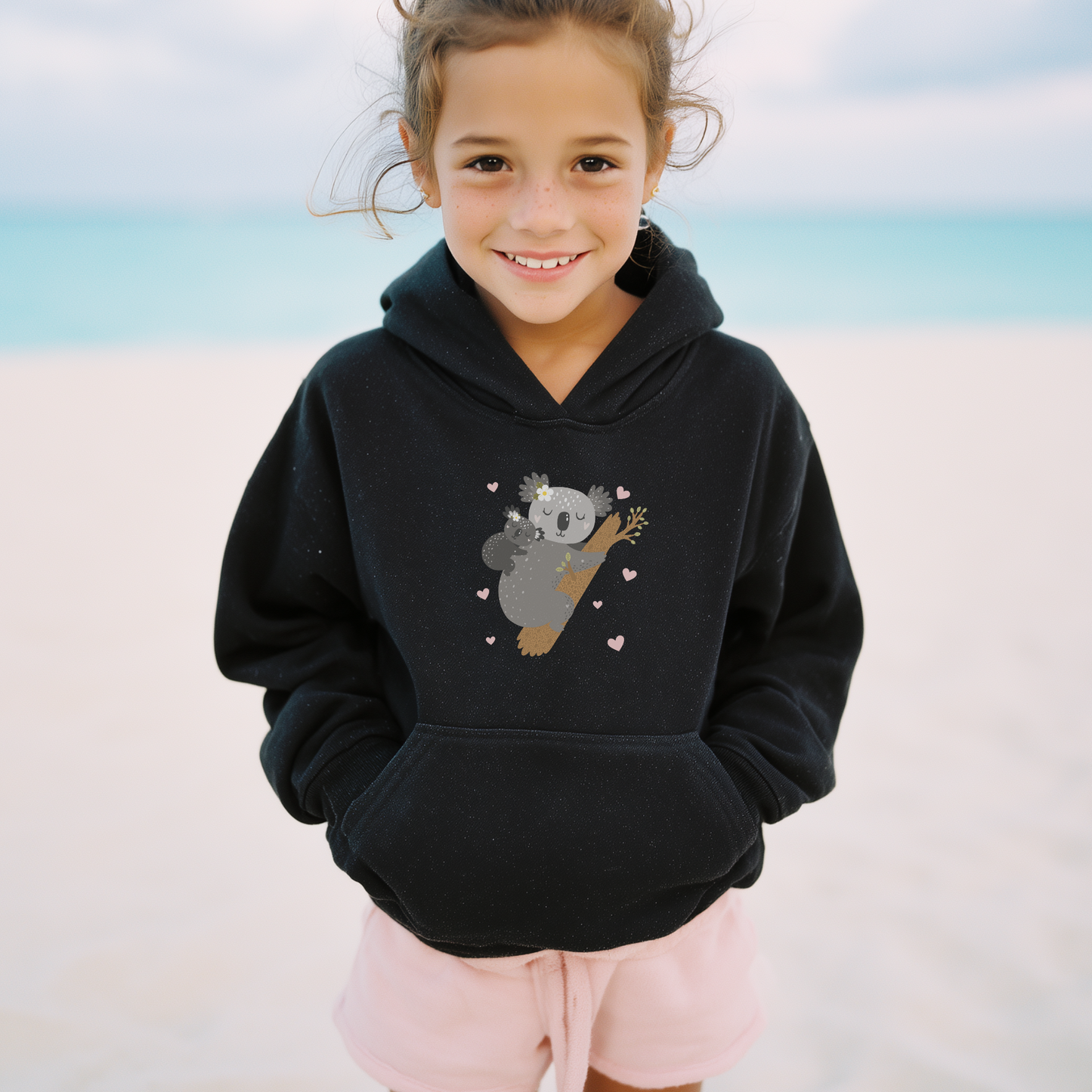 Young girl on a beach wearing a black hooded sweatshirt featuring an adorable illustration of a sleeping mother koala cuddling her baby on a tree branch, surrounded by small pink hearts.