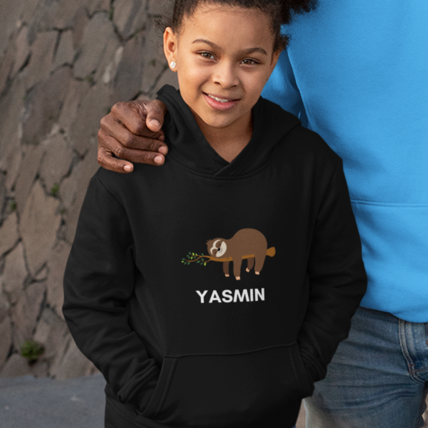 A smiling child with curly hair wears a black hooded sweatshirt featuring a cartoon sloth hanging on a branch and the a personalised name in white text. is printed below the sloth.
