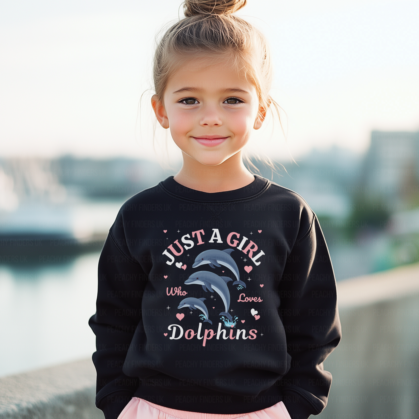 Young girl wearing a black sweatshirt with dolphin design and text by a waterfront.