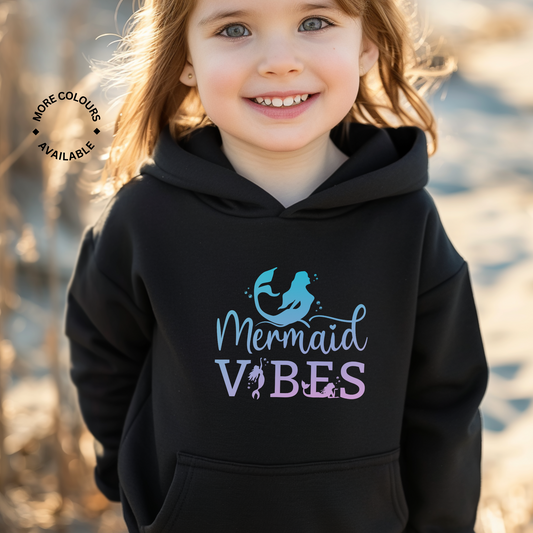 A little girl wearing a black hoodie with 'Mermaid Vibes' design standing on a beach