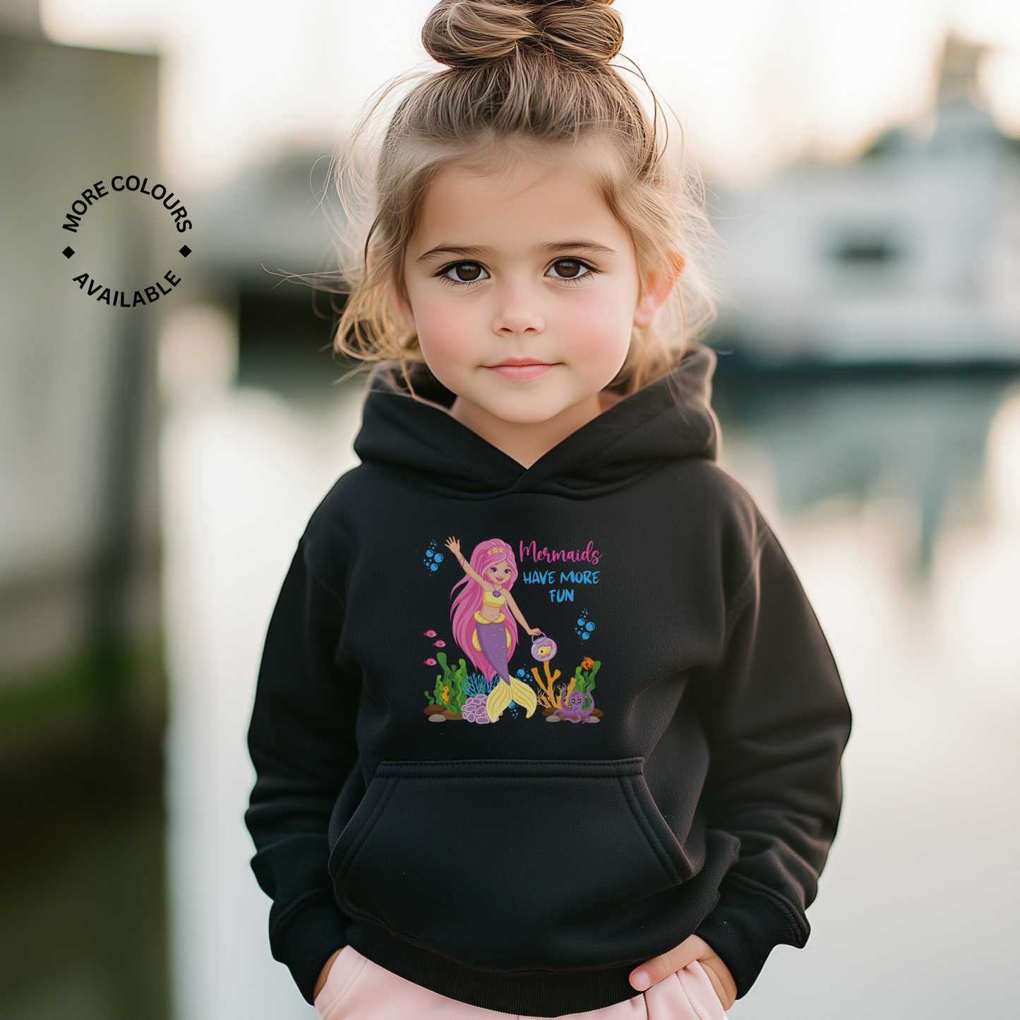 Young girl wearing a black hoodie with “Mermaids Have More Fun” text and colourful mermaid print on the front, smiling and posing outdoors.
