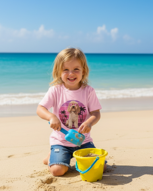 Little girl playing in the sand with a bucket on the beach wearing a pink children’s t-shirt featuring a summer cockapoo graphic and denim shorts