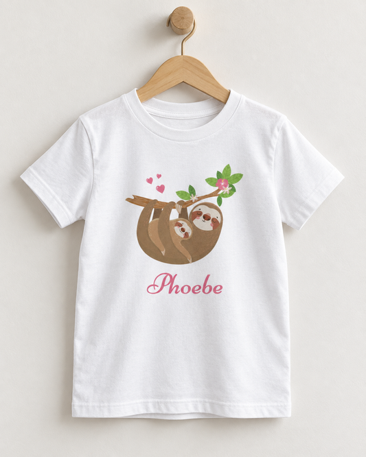 A white short-sleeve kids' t-shirt hanging on a wooden hanger against a light beige wall. The shirt features a cartoon graphic of a brown sloth hanging from a tree branch with green leaves and pink hearts, with the name 'Phoebe' printed below in pink cursive.