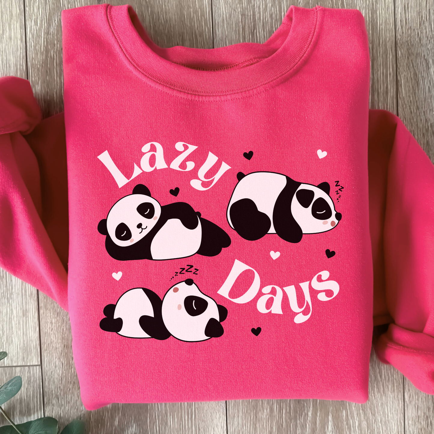 Girls pink sweatshirt with three pandas snoozing and the words “Lazy Days” surrounded by hearts, folded and styled on a wooden background