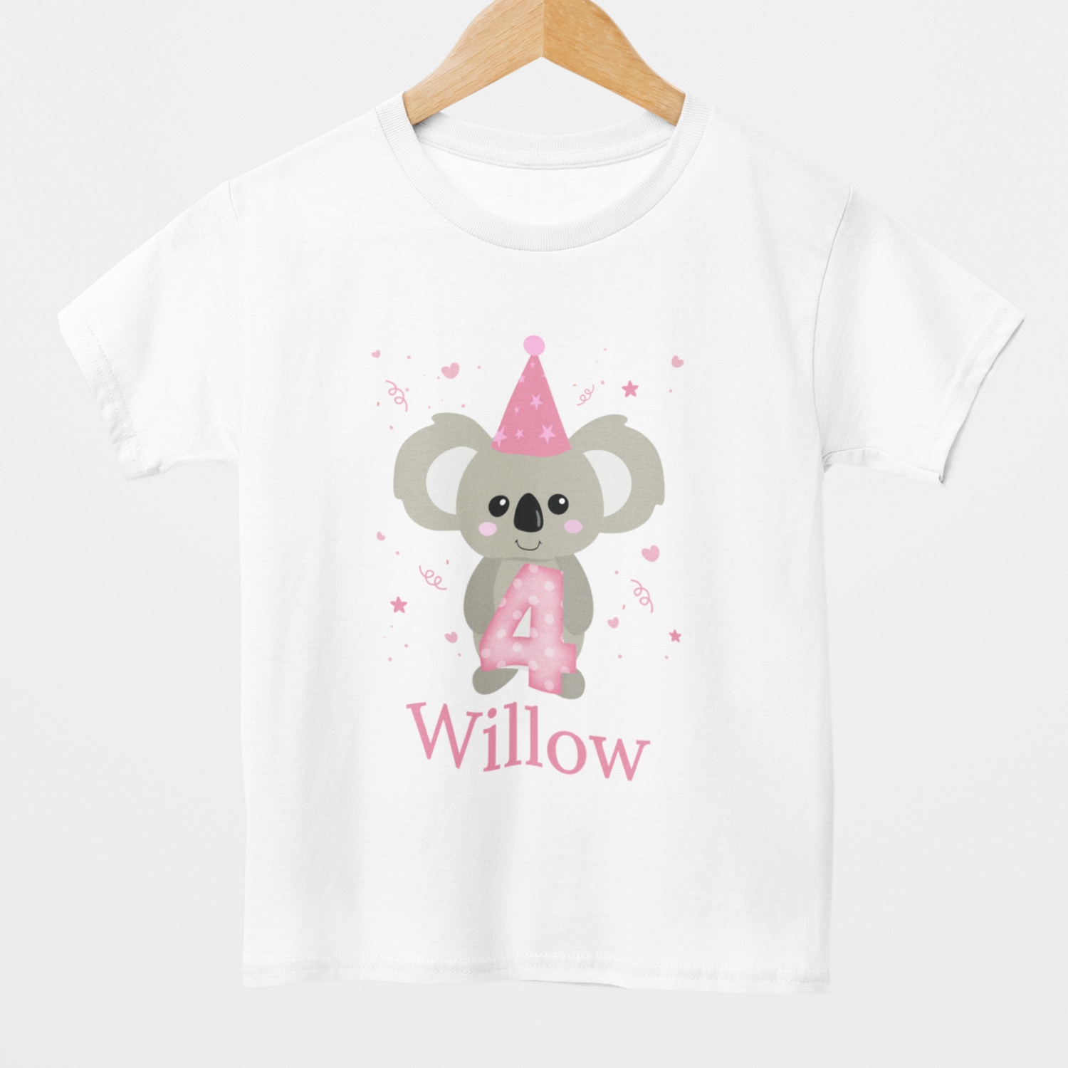 A kids white personalised birthday T-shirt styled on a wooden hanger, featuring a cute grey koala with a pink party hat, birthday number four with hearts and stars accents. Custom name printed underneath the design.