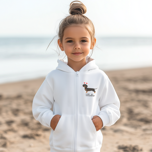 A young child smiles, wearing a white zip hoodie. The hoodie has a graphic of a black and tan dachshund with red hearts. The child's name, Mollie, is printed on the hoodie. The background is a blurred beach scene.