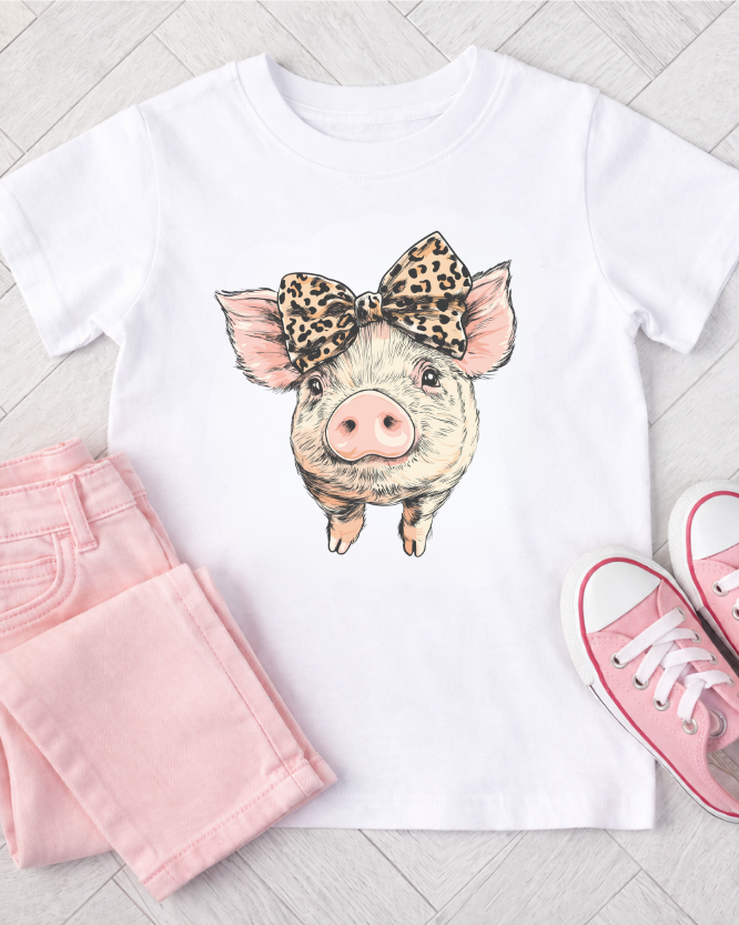 Kids white t-shirt with a pig graphic wearing a leopard print bow - styled with pink jeans, and pink sneakers on a light wooden floor.