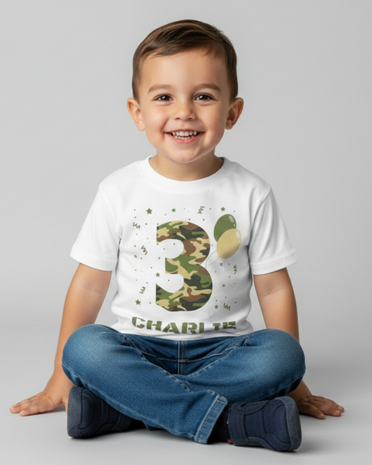 Happy child sitting cross-legged on the floor wearing a white personalised birthday T-shirt with camouflage number 3 and custom name, studio background.