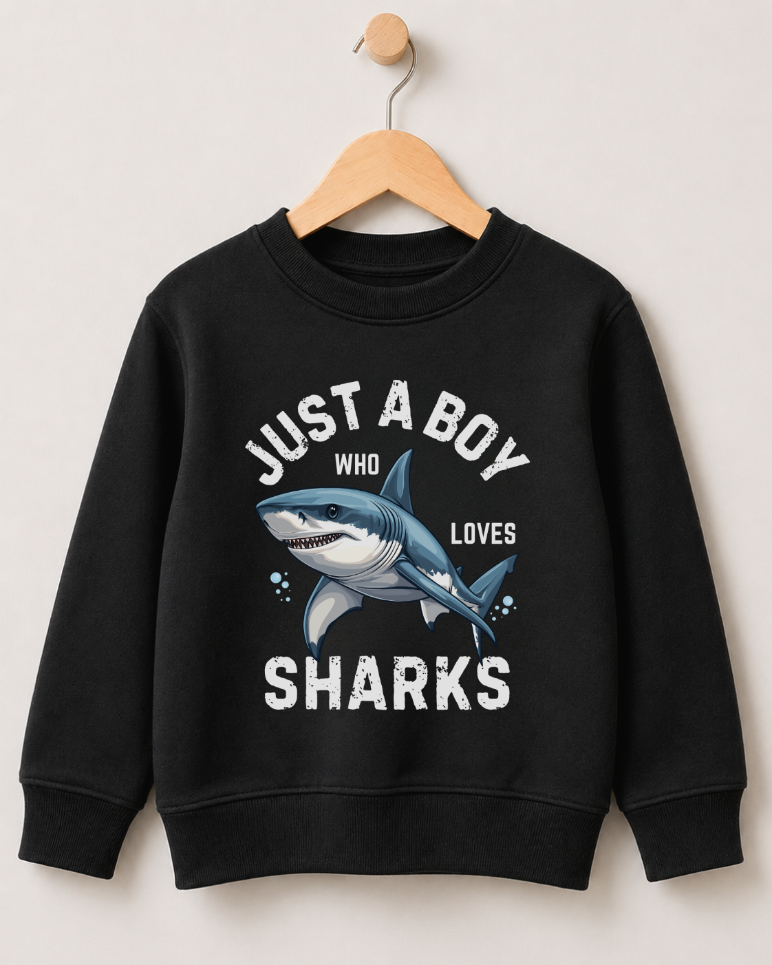 A kids black crewneck sweatshirt for kids hanging on a wooden hanger against a light beige wall. The sweatshirt features a graphic print of a blue and white shark with the text 'JUST A BOY WHO LOVES SHARKS' in white.