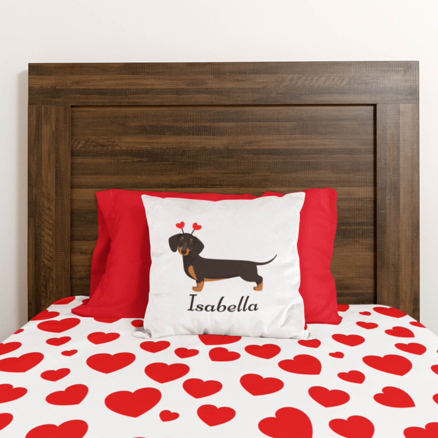 A white square cushion cover featuring a cartoon dachshund with heart-shaped antennae and the name "Isabella" printed below. The cushion is placed on a bed with a red and white heart-patterned bedspread.