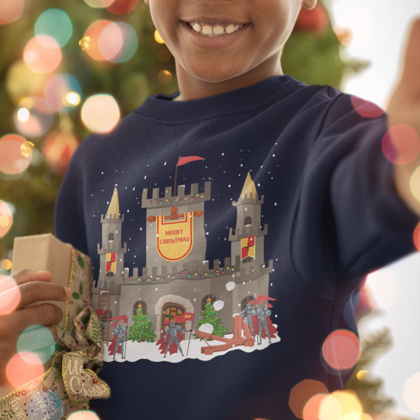 boy wears navy Christmas jumper featuring a snowy stone castle with “Merry Christmas” banner, golden turrets, red-armoured knights, catapulting snow-balls and festive trees on the front.