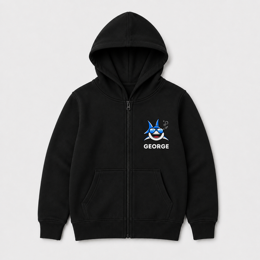 A kids black zip-up hoodie with a front kangaroo pocket, featuring a cartoon shark graphic and the name 'GEORGE' printed in white on the left chest, displayed on a light grey background.