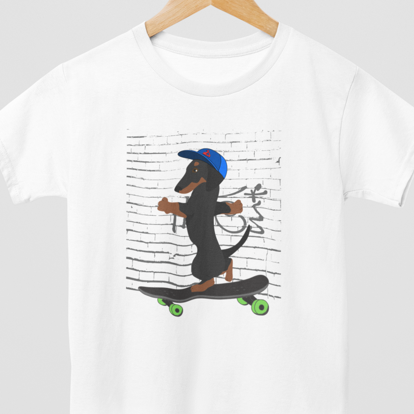 Kids white t-shirt on a wooden hanger, featuring a cool printed graphic of a Dachshund Dog wearing a baseball cap riding a skateboard with green wheels