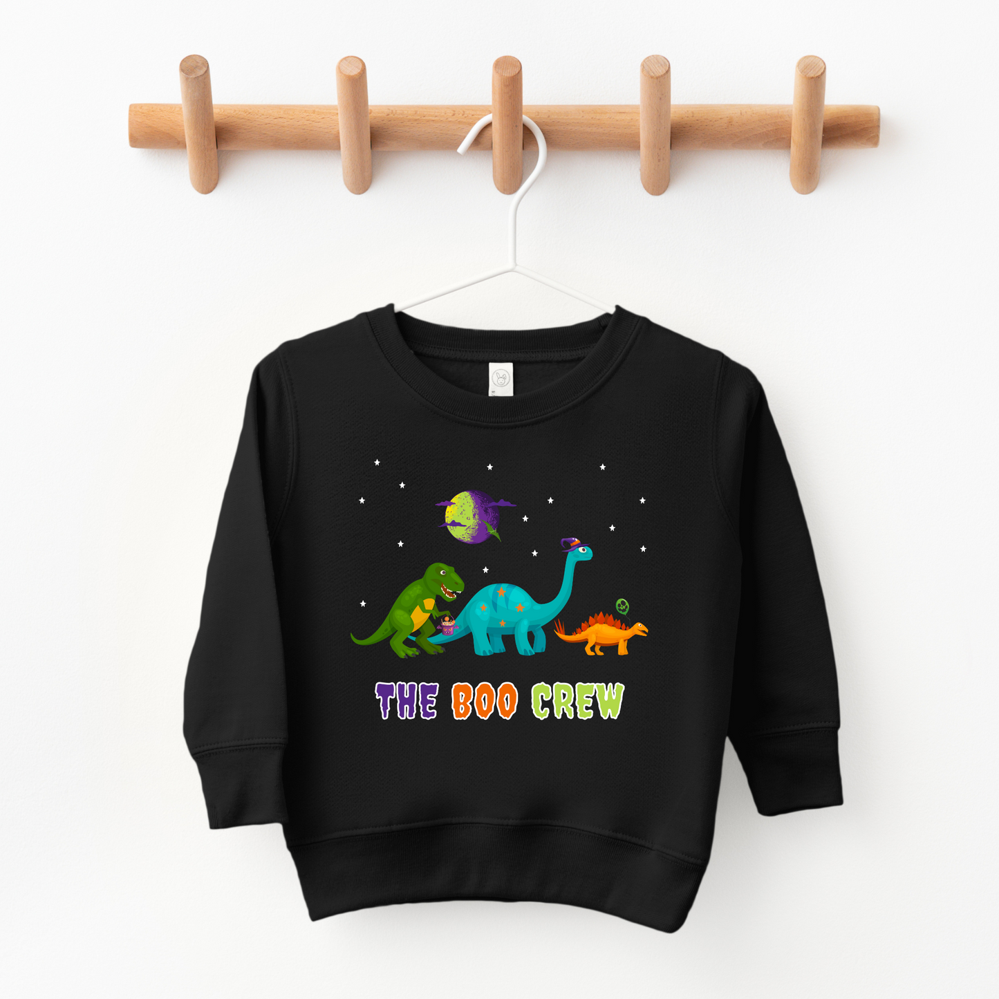 kids sweatshirt green Tyrannosaurus Rex holding a white trick-or-treat bucket filled with candy. In the centre is a teal Brontosaurus wearing a purple and blue striped hat. To the right is a small, orange Stegosaurus holding a green and orange patterned balloon. The dinosaurs appear to be walking or standing together as "THE BOO CREW".
