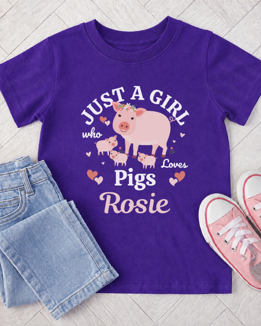 A kids purple short-sleeve t-shirt with a cartoon pig graphic and the text 'JUST A GIRL who Loves Pigs Rosie' in white and pink lettering. The shirt is styled in a flat lay with light blue jeans and pink sneakers on a light grey wood plank background.