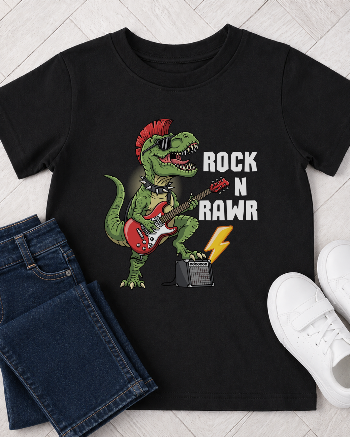 A kids black short-sleeve graphic T-shirt laid flat on a light wood floor, styled with folded blue jeans on the left and white sneakers on the right. The shirt features a green dinosaur playing a red electric guitar with the text 'ROCK N RAWR' in white.
