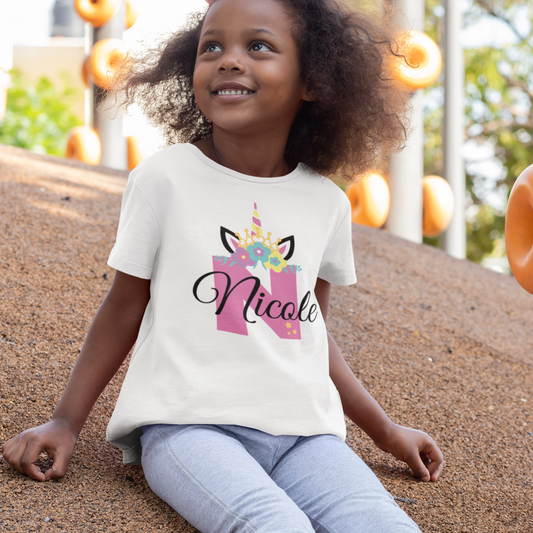 Little girl in a park wearing a white cotton t-shirt featuring a pink unicorn horn and ears design with colourful floral crown and stars. Personalised with a large initial and custom name text.