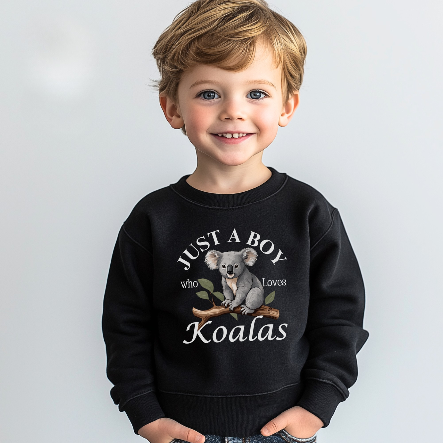 A little boy modelling a black sweatshirt featuring a printed design of a cute koala sitting on a brown tree branch with green leaves, surrounded by the words “Just A Boy Who Loves Koalas” in white text.