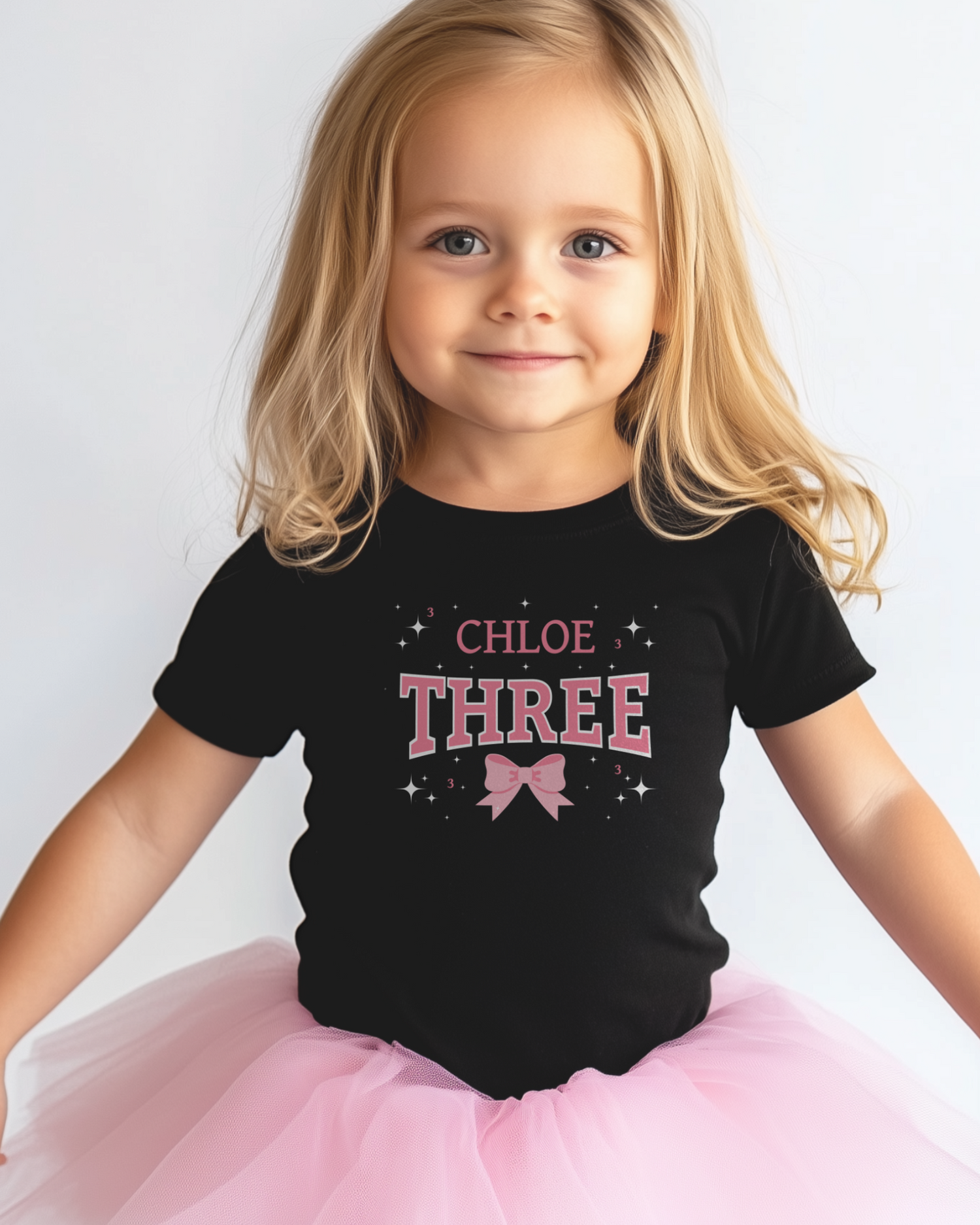 Young girl wearing a black personalised “Three” birthday T-shirt with pink bow graphic and a fluffy pink tutu, smiling against a plain light background.