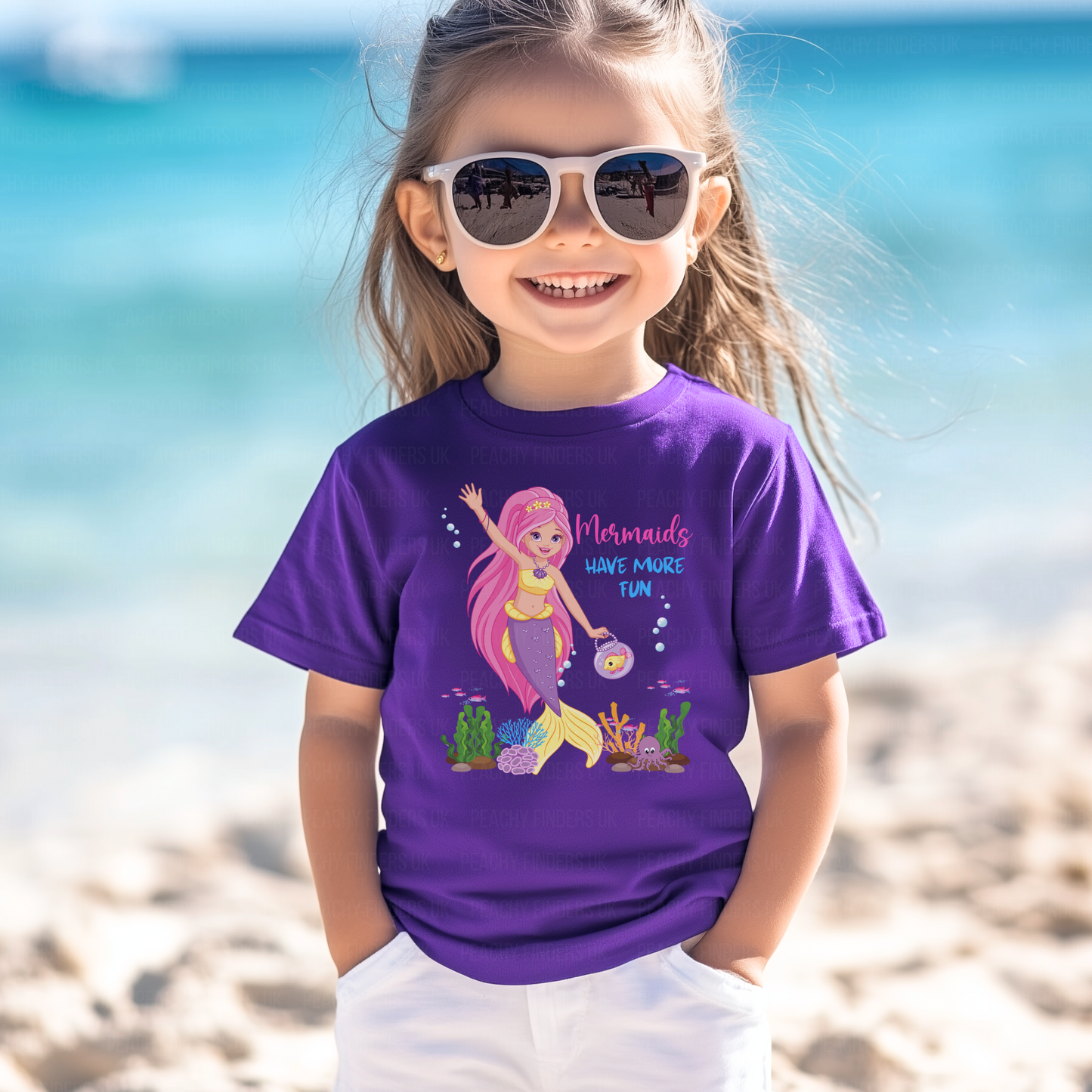 A young child with light brown hair smiles while wearing white sunglasses and a purple t-shirt. The t-shirt features a colourful cartoon mermaid graphic with pink hair and a purple and yellow tail, along with the text "Mermaids HAVE MORE FUN". The background is a bright, blurred beach scene with light sand and turquoise sea.