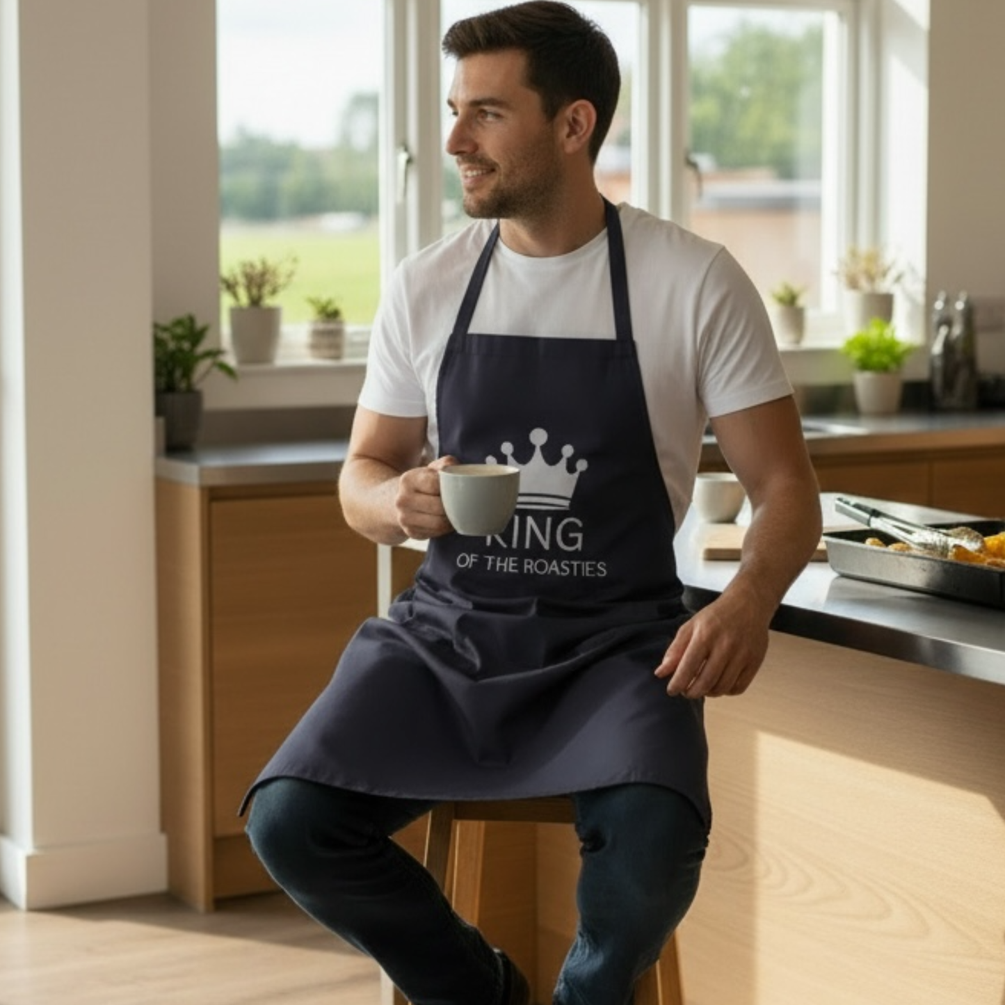 Man wearing navy “King of the Roasties” kitchen apron sitting on a stool, novelty cooking apron for dads and roast dinner lovers