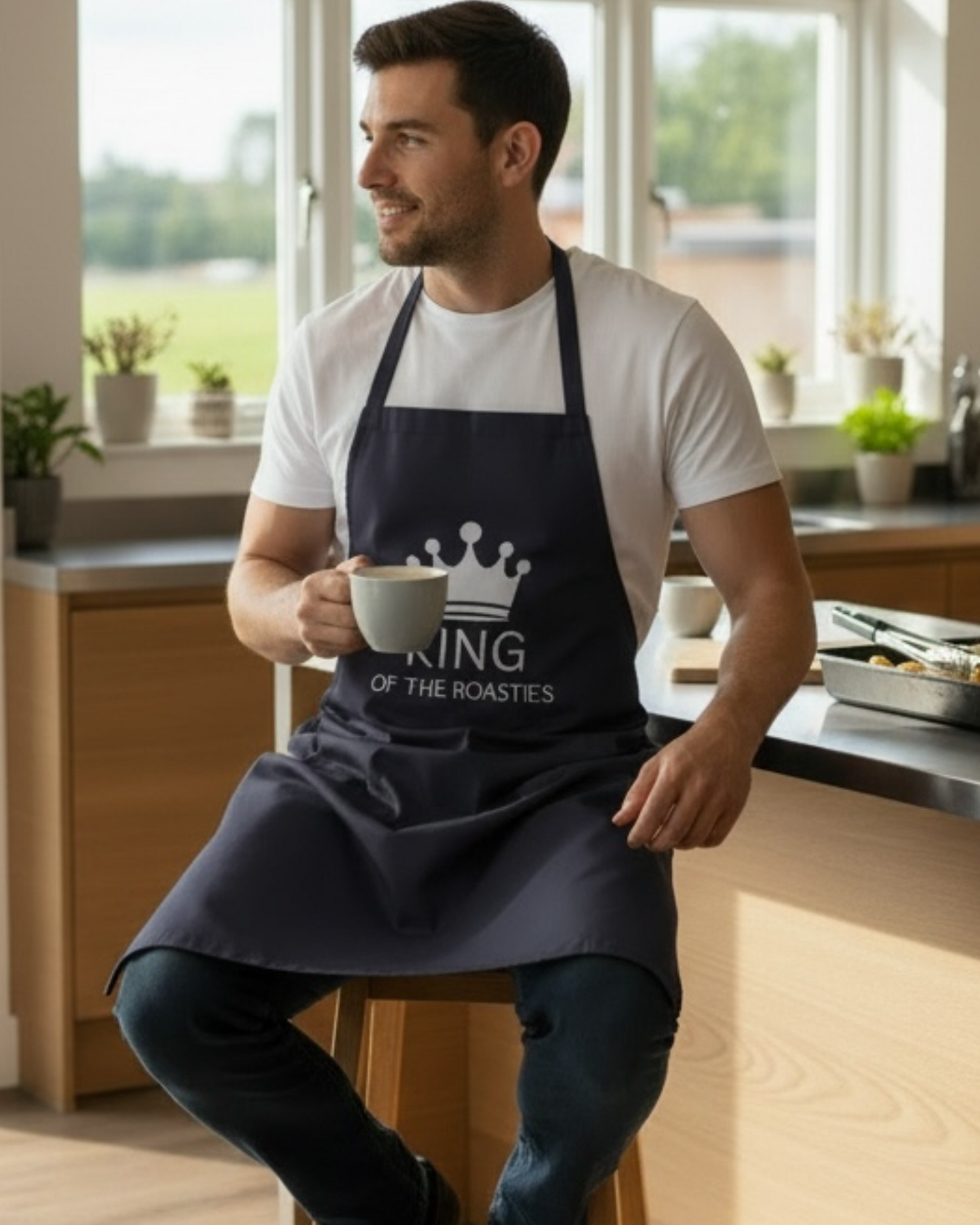 Man wearing navy “King of the Roasties” kitchen apron sitting on a stool, novelty cooking apron for dads and roast dinner lovers
