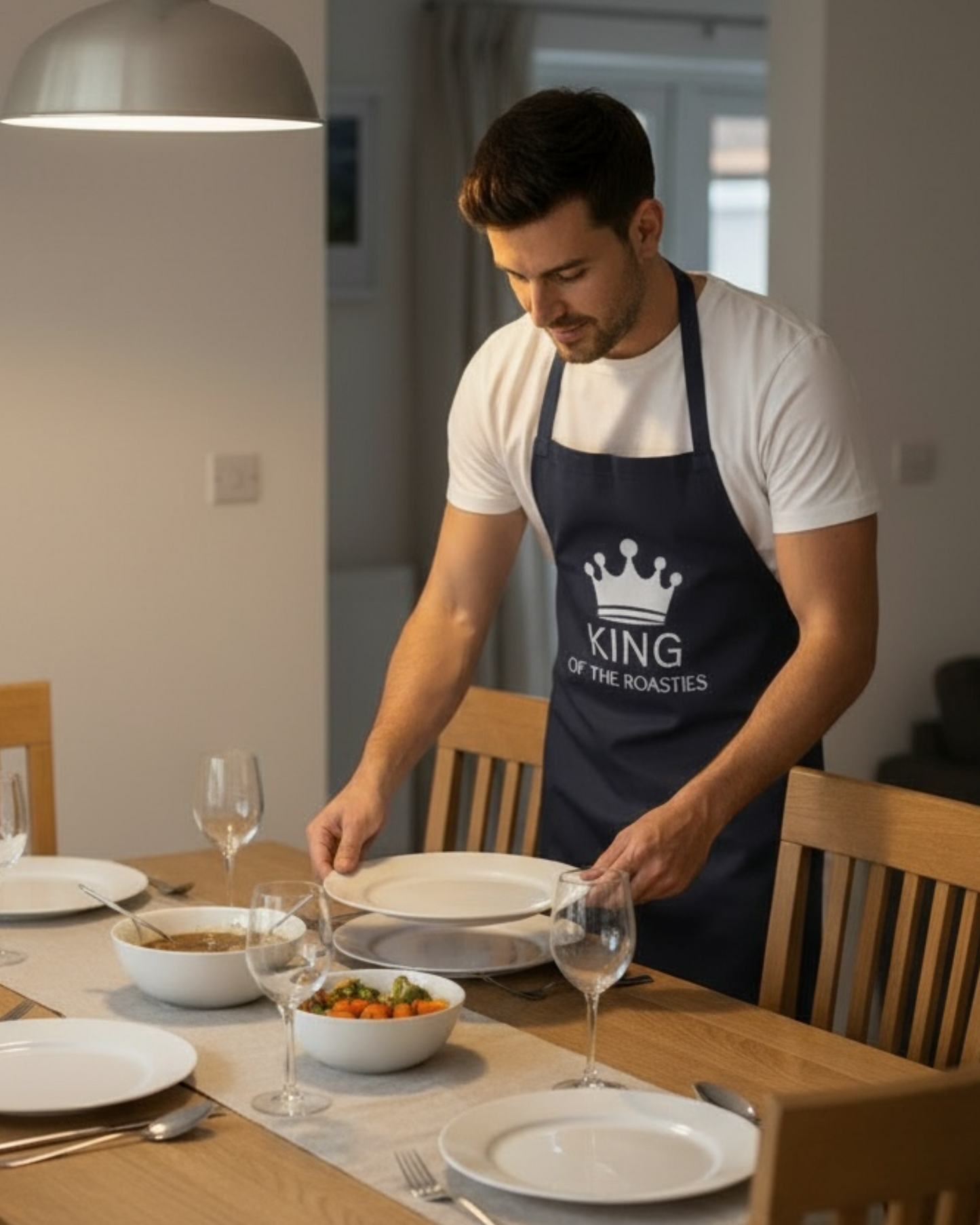 Man laying a dining table while wearing a navy King of the Roasties apron, fun gift apron for home cooks and Sunday roast chefs
