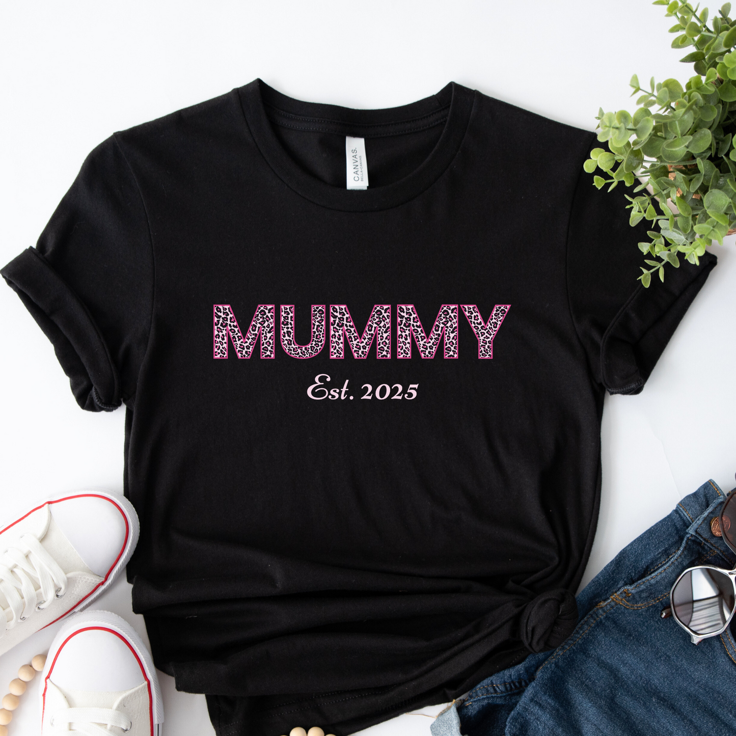 Black t-shirt with the word “MUMMY” in pink leopard print and “Est. 2025” underneath, laid flat next to white trainers, denim shorts, and sunglasses.