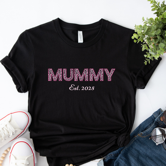 Black t-shirt with the word “MUMMY” in pink leopard print and “Est. 2025” underneath, laid flat next to white trainers, denim shorts, and sunglasses.