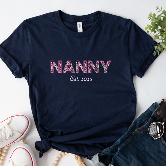 Flat lay of a navy t-shirt with "NANNY" in pink leopard print and "Est. 2025" underneath. Styled with white trainers and denim shorts.