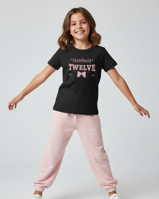 Girl aged 12 wearing black Hannah Twelve birthday shirt with pink bow design, standing in studio with pink joggers and white trainers