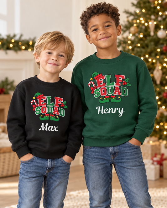 Two smiling young boys posing together indoors near a Christmas tree, wearing matching personalised Christmas sweatshirts featuring an "ELF SQUAD" graphic with candy canes, elf shoes, and their names "Max" and "Henry" printed underneath.